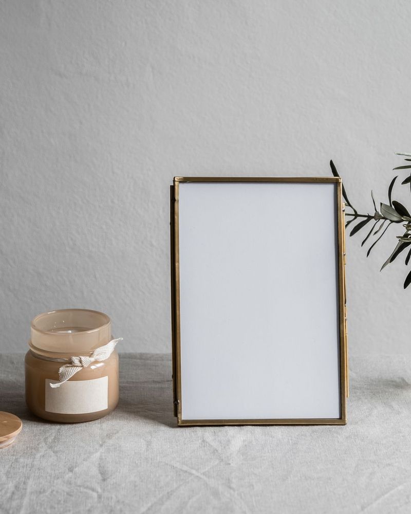 A brass-edged photo frame on linen beside a candle and olive sprig
